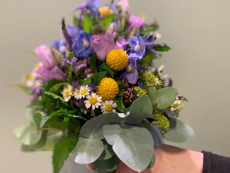 Colorful bouquet of purple flowers, yellow button mums, white daisies, and green foliage tied with a gray ribbon