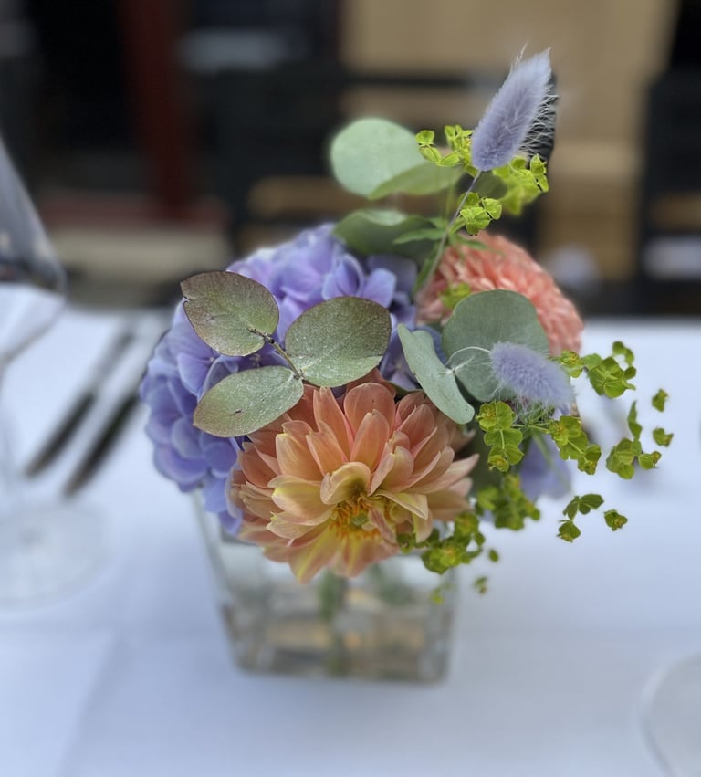 A small floral arrangement in a clear vase featuring peach dahlias, purple flowers, eucalyptus leaves, and ornamental greenery on a white table setting