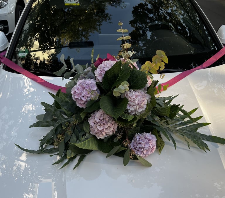 Floral wedding car decoration featuring pink hydrangeas and yellow flowers with pink ribbons on white car hood