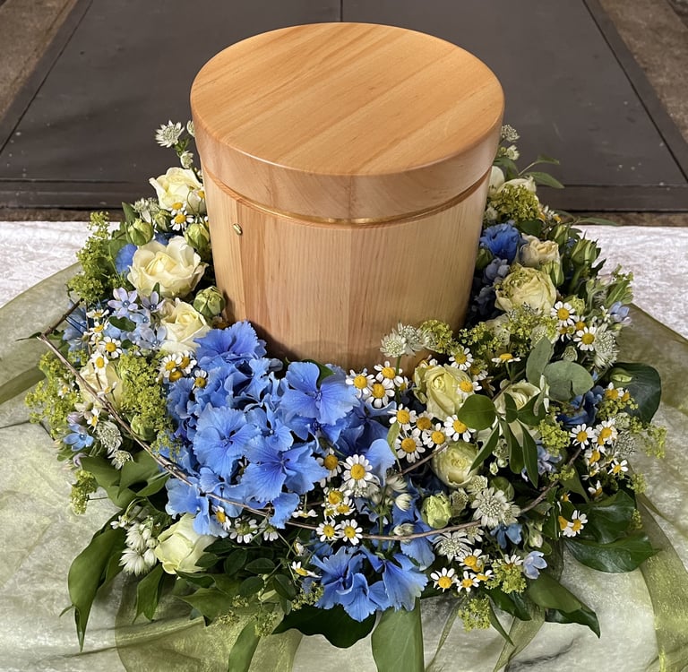 Wooden cremation urn surrounded by a floral wreath of blue and white flowers with green foliage