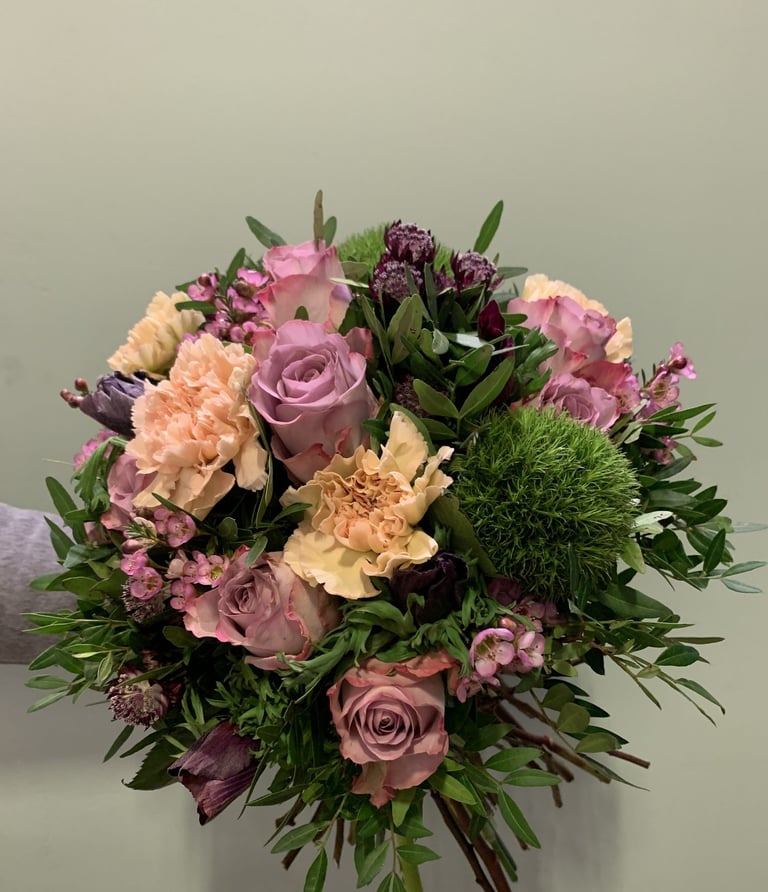 Lush floral bouquet with pink roses, mauve carnations, peach flowers, and burgundy blooms arranged with green foliage against neutral background