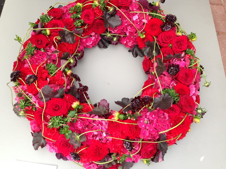 Vibrant floral wreath with red and pink flowers, dark foliage, and greenery arranged in a circular ring on white background
