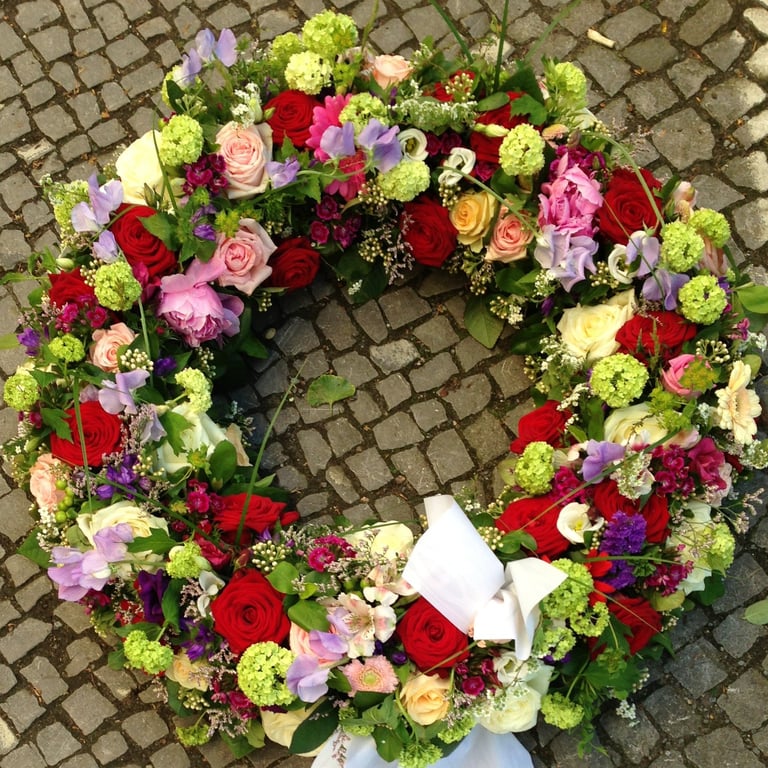 Vibrant flower wreath with red roses, pink carnations, purple flowers, and green chrysanthemums arranged on a brick surface with a white bow