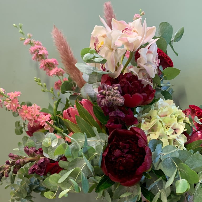 Lush mixed flower bouquet featuring dark red peonies, pink roses, pale orchids, cream hydrangeas, and decorative greenery against a soft sage background