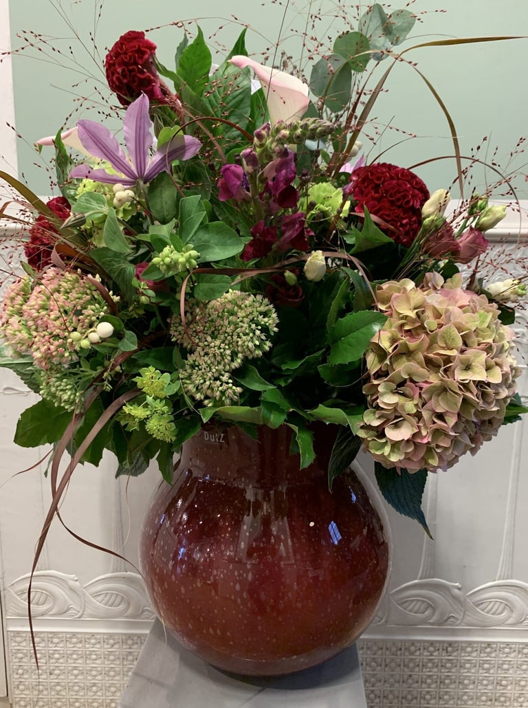 Floral arrangement in burgundy vase with red carnations, purple flowers, green hydrangeas, and dried foliage
