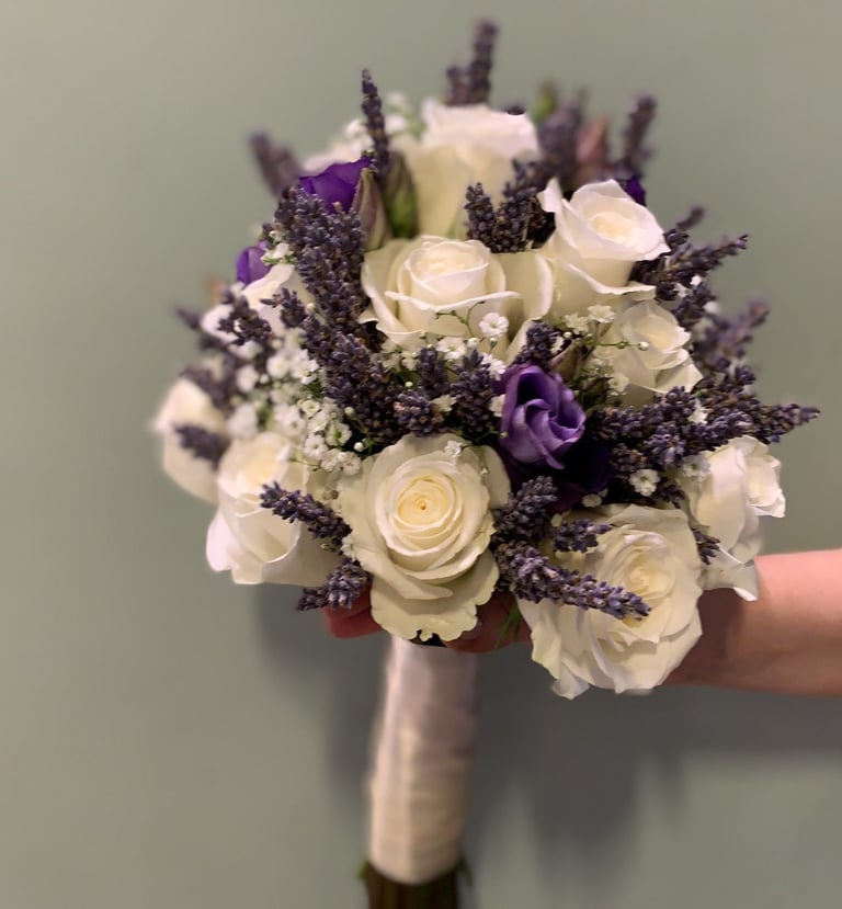 Bridal bouquet with white roses, purple flowers, lavender sprigs, and greenery wrapped in silver ribbon and cream-colored handle.