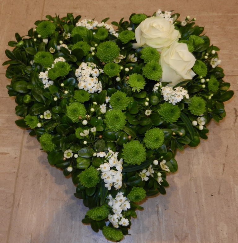 Heart-shaped funeral flower arrangement with white roses, green chrysanthemums, and small white flowers on wooden floor