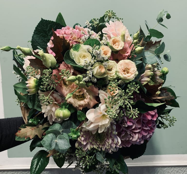 Lush bouquet of pink, cream, and burgundy flowers with greenery held by a person in black against a gray background